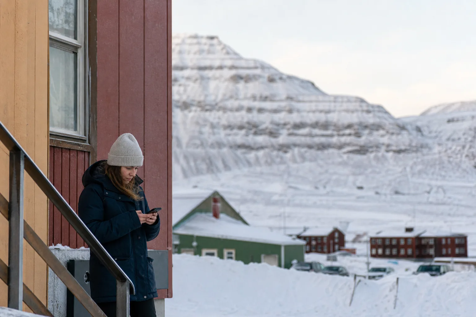 Snötäckt gruvstad i Norrbotten med kala fjäll i bakgrunden och en person i vinterkläder som tittar på mobilen utanför ett bostadshus i kallt midvinterljus