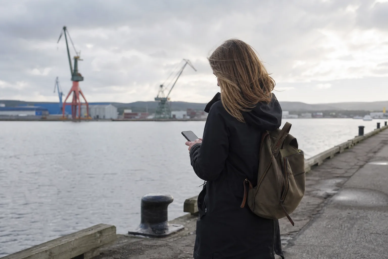Industristad vid en fjärd i norra Sverige med hamnkranar och hav och en person med mobil på kajpromenaden en mulen dag