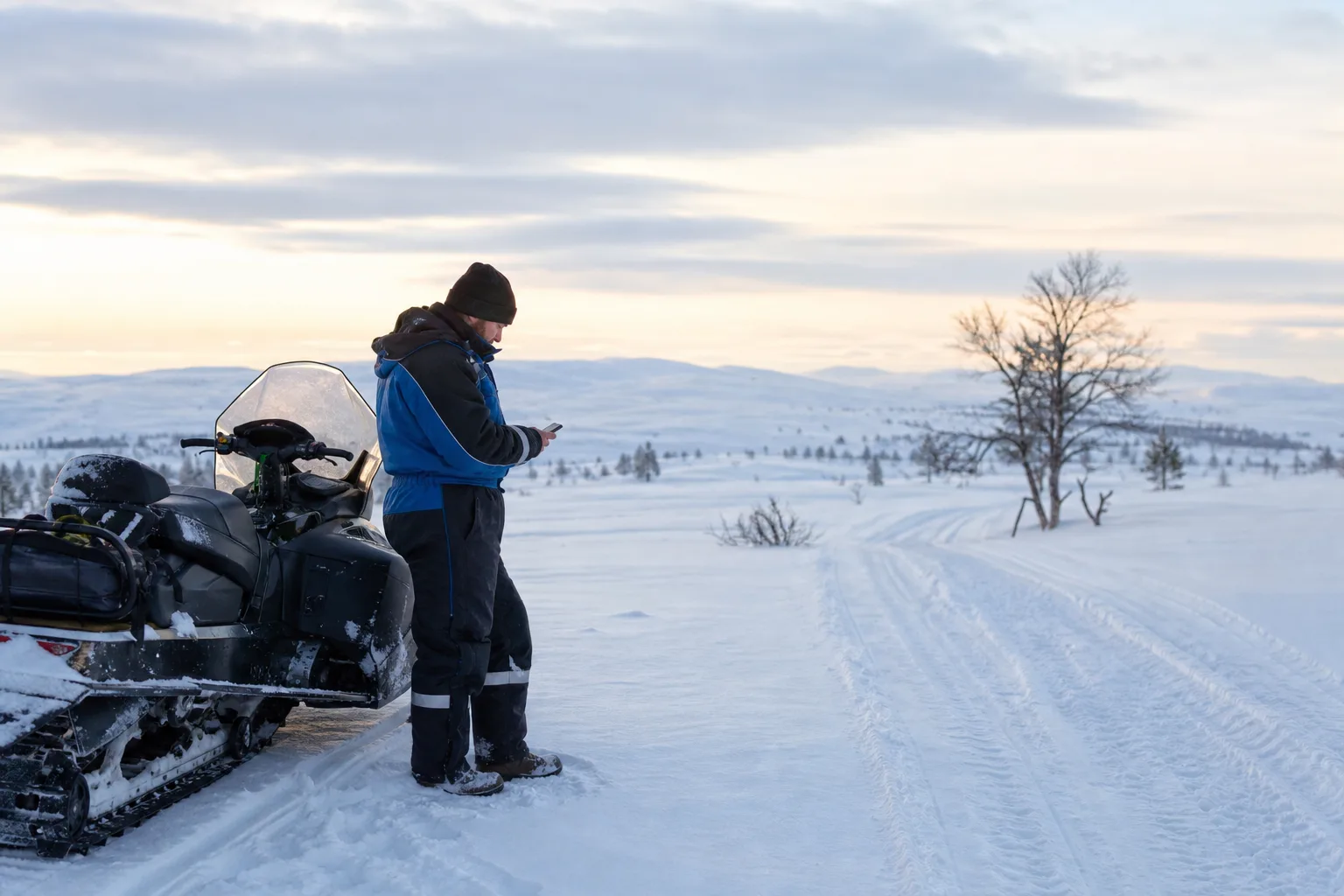 Snöskoterförare som stannat på en led i fjällterräng och tittar på mobilen i ett vidsträckt vitt vinterlandskap