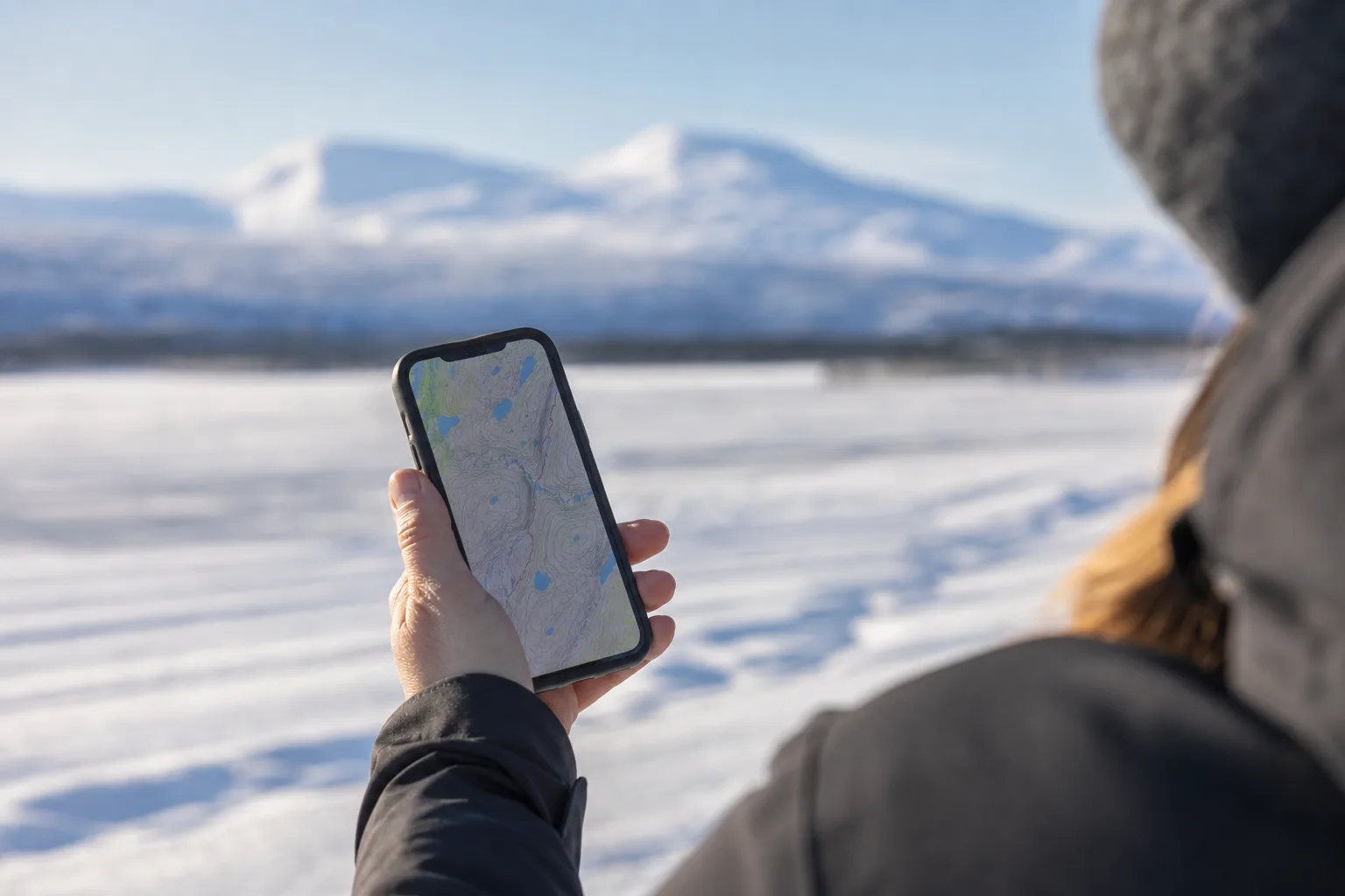 Hand som håller en mobil med täckningskartan öppen i ett snötäckt landskap i norra Sverige med suddiga fjäll i bakgrunden