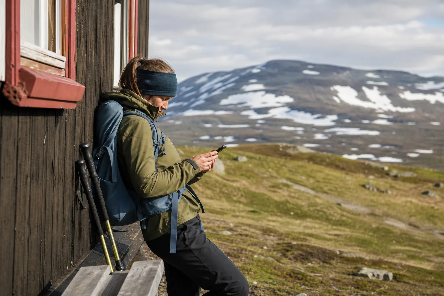 Vandrare kollar mobilen vid en fjällstuga i norra Sverige, med snöfläckar och kala fjäll – Telias nät når längst i fjällterräng.
